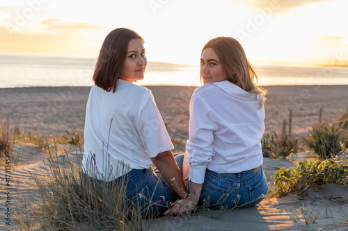 Joven pareja gay disfrutando de unas vistas increíbles del atardecer en el mar sentadas y abrazadas vestidas con camisa blanca y vaqueros.