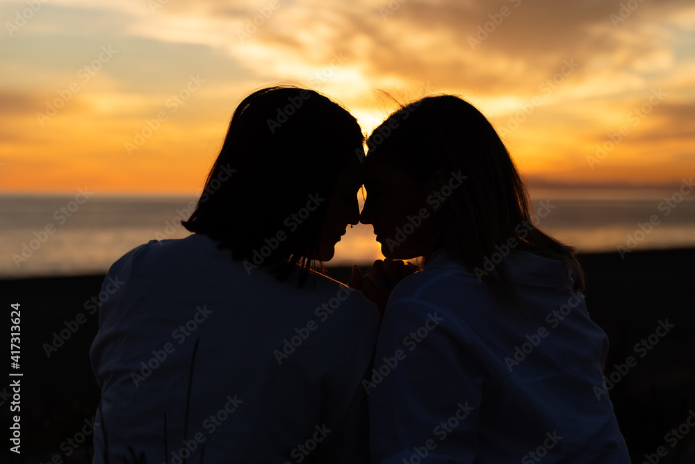 Joven pareja lesbiana mirándose bajo un atardecer a través de una silueta oscura con precioso mar y fondo de nubes con sol