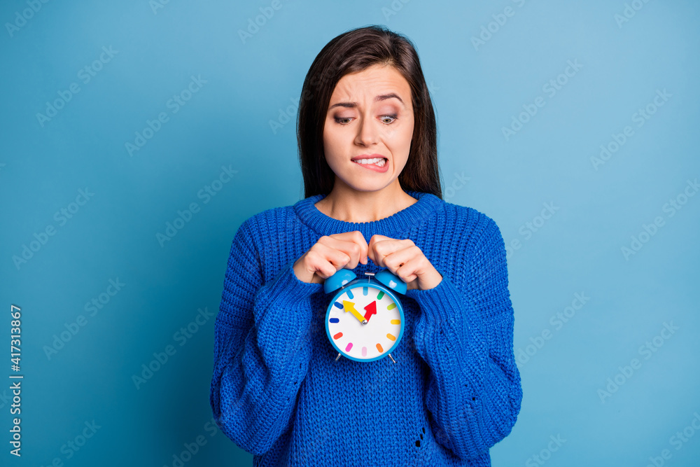 Photo of unhappy upset sad puzzled nervous girl bite lips hold clock wear knitted sweater isolated on blue color background Photo of unhappy upset sad puzzled nervous girl bite lips hold clock wear knitted sweater isolated on blue color background