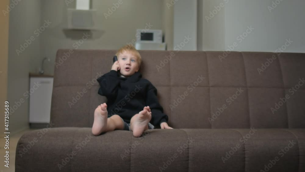 Happy little boy in black hoodie sits on couch and talks on phone with mother Happy little boy in black hoodie sits on couch and talks on phone with mother