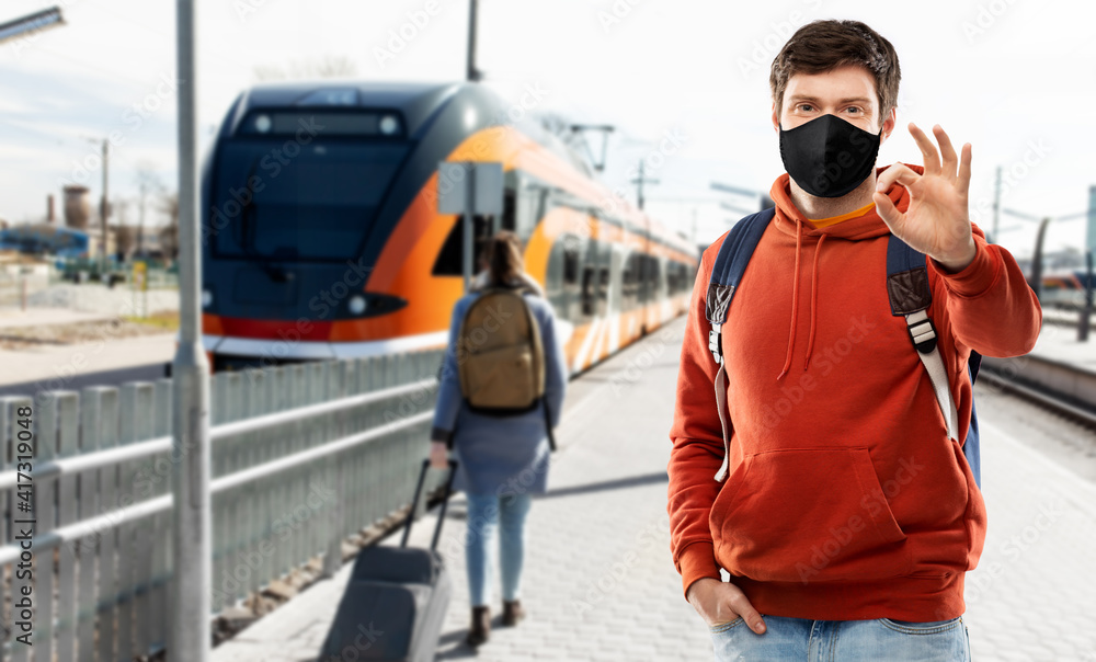 travel, tourism and pandemic concept - young man with backpack in face protective medical mask showing ok hand sign traveling by train over railway station in city of tallinn, estonia on background travel, tourism and pandemic concept - young man with backpack in face protective medical mask showing ok hand sign traveling by train over railway station in city of tallinn, estonia on background