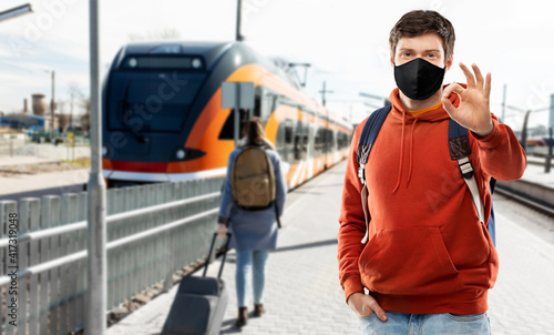 travel, tourism and pandemic concept - young man with backpack in face protective medical mask showing ok hand sign traveling by train over railway station in city of tallinn, estonia on background