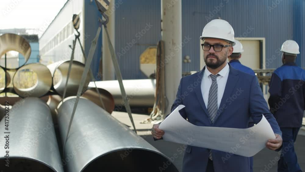 Slowmo PAN of serious male manufacturing engineer in suit and hard hat standing outdoors beside steel pipes and looking at blueprint. Workers in uniforms walking in background Slowmo PAN of serious male manufacturing engineer in suit and hard hat standing outdoors beside steel pipes and looking at blueprint. Workers in uniforms walking in background