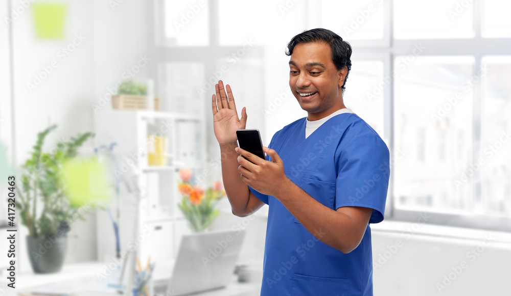 medicine, healthcare and technology concept - happy smiling indian doctor or male nurse in blue uniform having video call on smartphone over medical office at hospital background