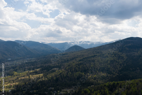 Summer mountain landscape, beautiful scenery of Carpathian mountains in Western Ukraine. High mountain peaks covered with snow, cloudy sky, green forests and a small village at the foot of the hill.