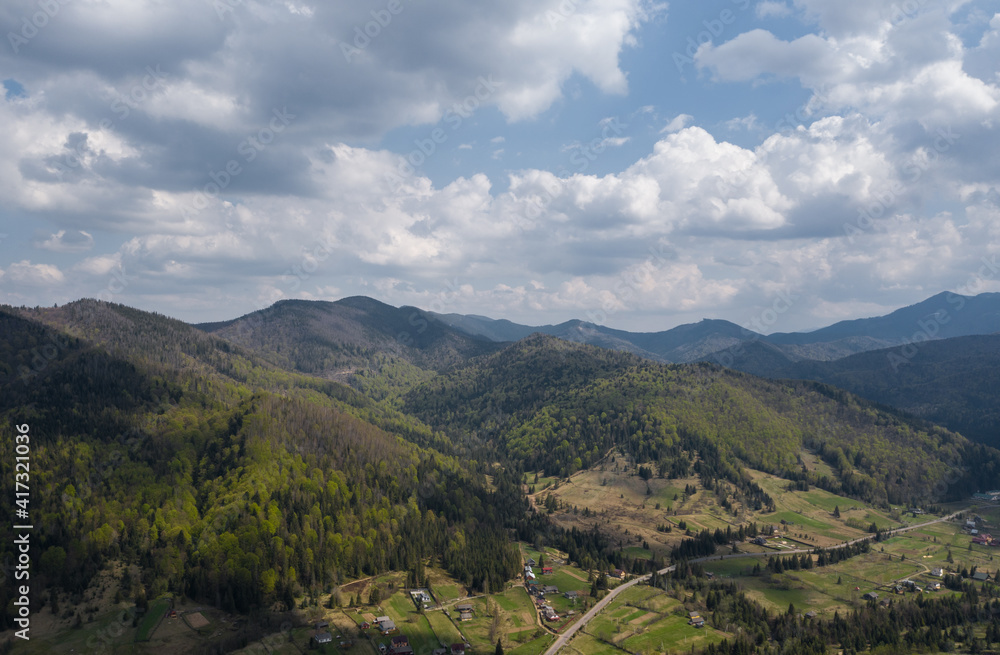 Summer mountain landscape, beautiful scenery of Carpathian mountains in Western Ukraine. High mountain peaks, cloudy sky, green forests and a small village at the foot of the hill. Panoramic view.