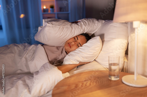 people, bedtime and rest concept - annoyed african american woman lying in bed at night and covering ears with pillow