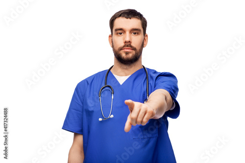healthcare, profession and medicine concept - doctor or male nurse in blue uniform with stethoscope pointing finger to camera over white background