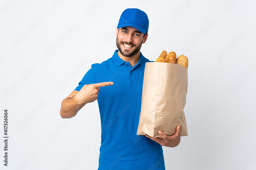 Delivery man holding a bag full of breads isolated on white background and pointing it Delivery man holding a bag full of breads isolated on white background and pointing it