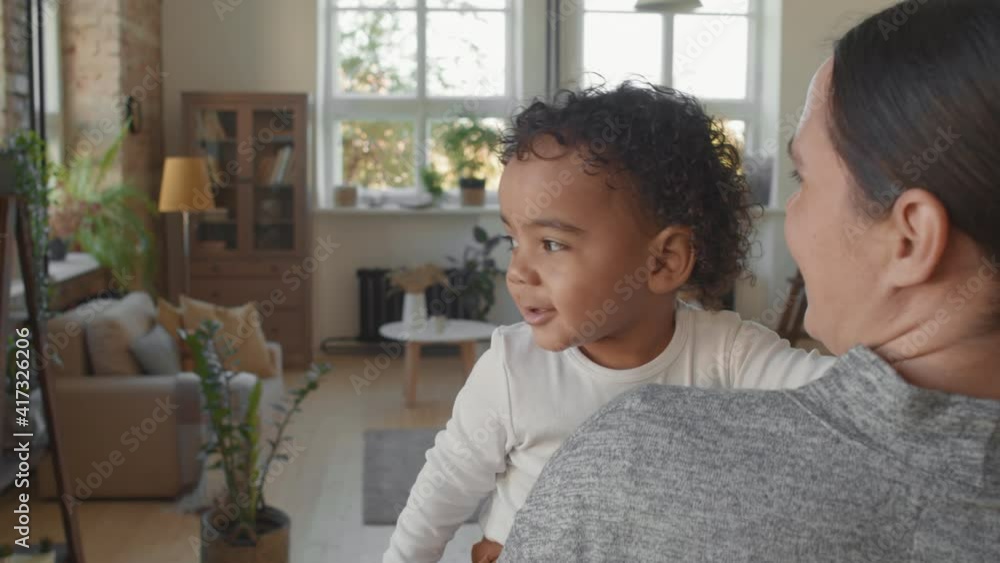 Slowmo shot of cheerful mother holding adorable black toddler boy and talking to him while he is looking out window with curiosity Slowmo shot of cheerful mother holding adorable black toddler boy and talking to him while he is looking out window with curiosity