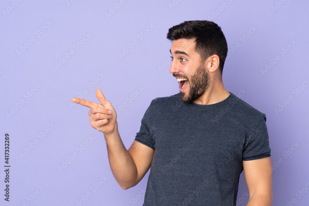 Caucasian handsome man pointing finger to the side and presenting a product over isolated purple background Caucasian handsome man pointing finger to the side and presenting a product over isolated purple background