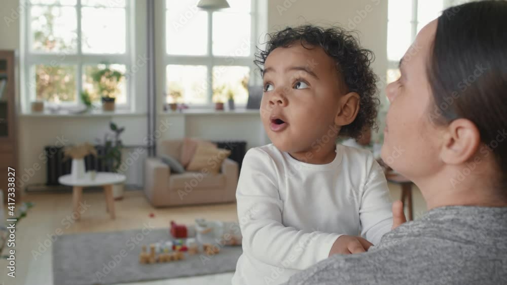 Slowmo shot of happy mother standing by window in cozy loft apartment and holding adorable toddler boy. She is talking to him while he is curiously looking around Slowmo shot of happy mother standing by window in cozy loft apartment and holding adorable toddler boy. She is talking to him while he is curiously looking around
