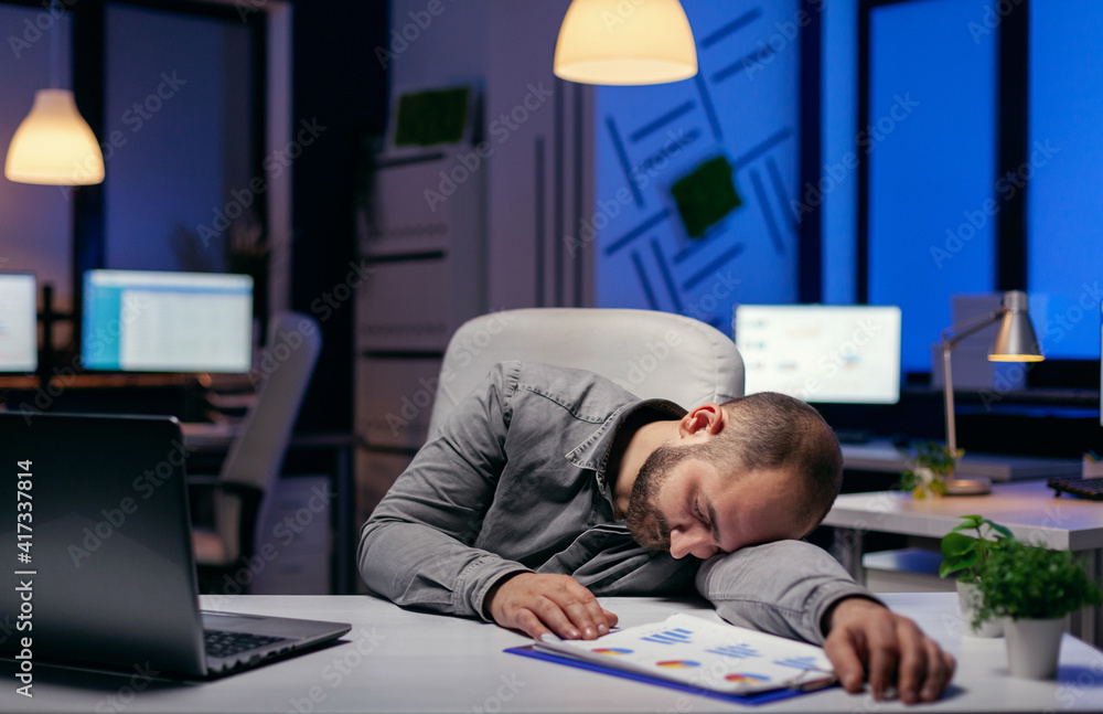 Tired businessman sleeping in his workplace on desk. Workaholic employee falling asleep because of while working late at night alone in the office for important company project. Tired businessman sleeping in his workplace on desk. Workaholic employee falling asleep because of while working late at night alone in the office for important company project.