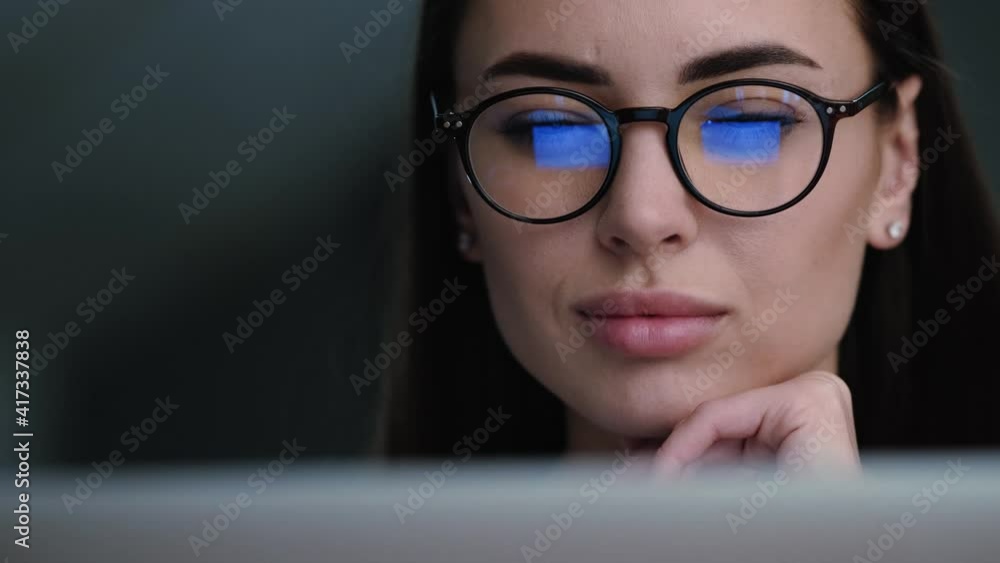 A close-up view of a concentrated young woman wearing glasses is using her laptop while sitting at the apartments A close-up view of a concentrated young woman wearing glasses is using her laptop while sitting at the apartments