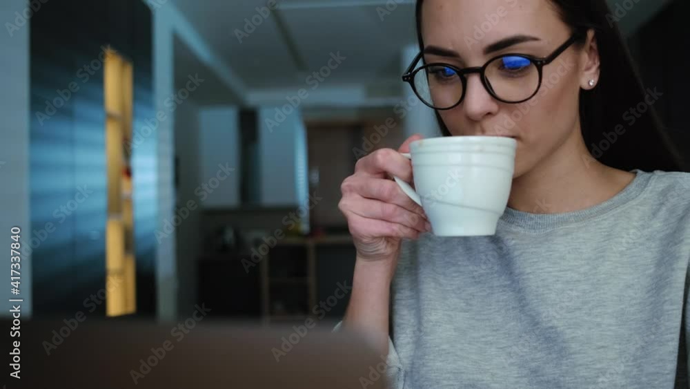 An attractive young woman wearing glasses is using her laptop while drinking tea sitting at the apartments An attractive young woman wearing glasses is using her laptop while drinking tea sitting at the apartments