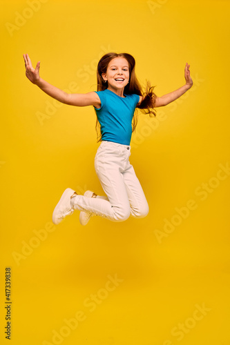 Positive girl wearing a blue t-shirt and white jeans, jumping with raised hands, on yellow background