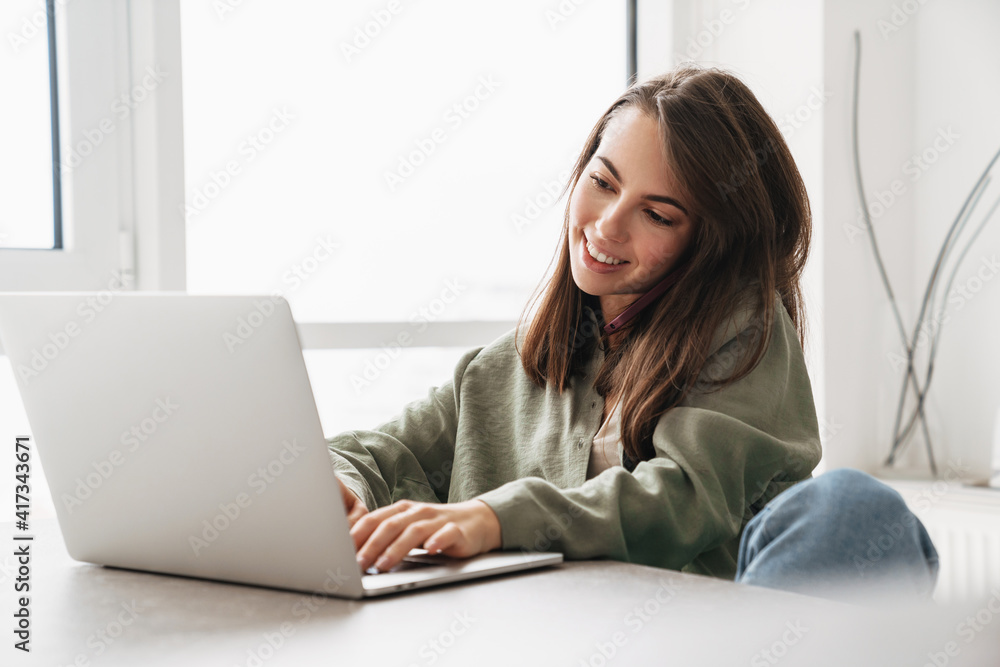 Fototapeta premium Smiling woman talking on cellphone while working with laptop