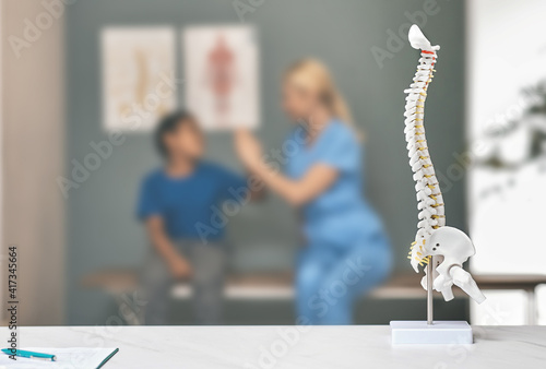 Anatomical model of spine on therapist's table in doctor's office. Boy patient and physiotherapist during a consultation on background, soft focus