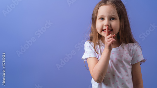 portrait of a cute little girl, holding her index finger to her mouth, stands on a purple background. The child doesn't want to talk. Keeps a secret, learned a secret