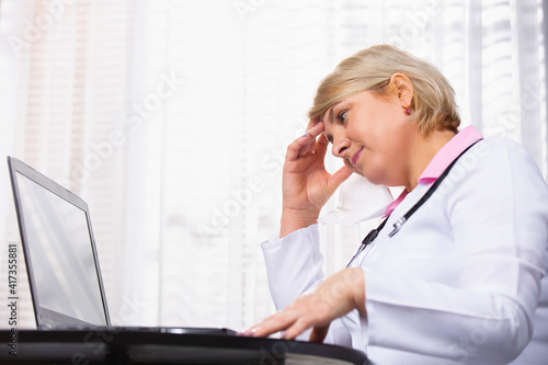 Senior woman doctor at the computer in her office holding her head. Fatigue. Stress. Work days.