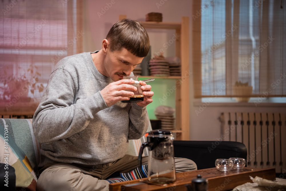 Young man tasting a freshly brewed cup of authentic chinese tea from a glass bowl. Bamboo tray and tea set on the table Young man tasting a freshly brewed cup of authentic chinese tea from a glass bowl. Bamboo tray and tea set on the table