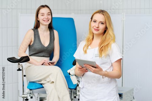 Принти на полотні a young female gynecologist doctor with a tablet in her hands against the background of a female patient sitting in a blue gynecological chair and a colposcope