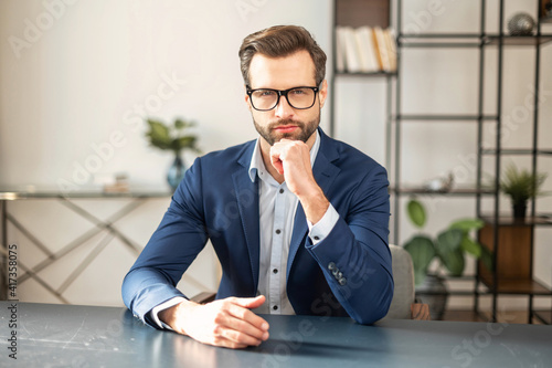 Portrait of sophisticated young bearded confident entrepreneur man in business casual clothes sitting at the desk and looking at camera, placed hand on his chin and thinking. Copy space concept photo