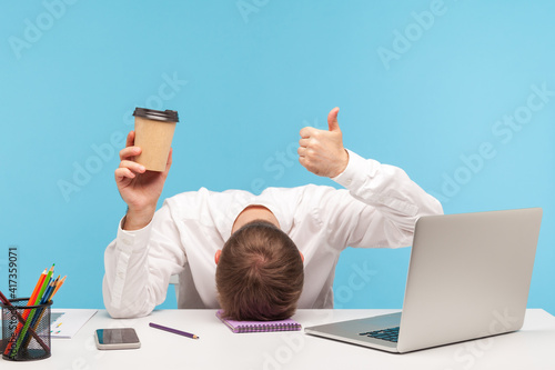 Bilde på lerret Tired man office worker lying face down on desk holding paper cup of coffee and showing thumbs up, need coffee to be ok, overworking