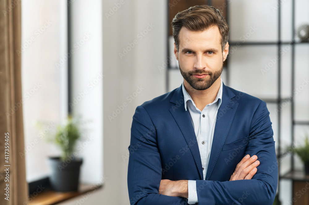 Successful serious authoritative confident young bearded brunet adult male entrepreneur businessman professional in formal wear standing with arms crossed at the modern home office, looking at camera Successful serious authoritative confident young bearded brunet adult male entrepreneur businessman professional in formal wear standing with arms crossed at the modern home office, looking at camera