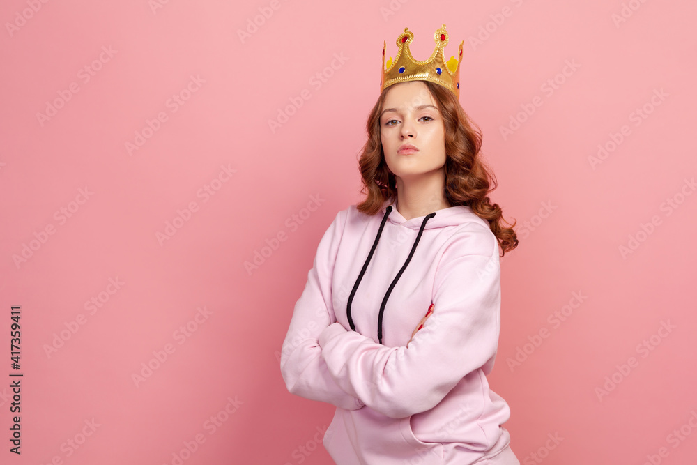 Portrait of confident curly haired teenage girl in hoodie with gold crown on head crossing hands and looking at camera, I am queen. Indoor studio shot isolated on pink background Portrait of confident curly haired teenage girl in hoodie with gold crown on head crossing hands and looking at camera, I am queen. Indoor studio shot isolated on pink background