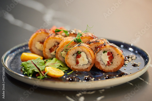 A close-up photo of sushi rolls with salmon, Philadelphia cheese, and tuna on a blue ceramic plate with sauce, sesame, lobules of kumquat, and leaves. Black stone table.