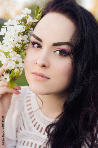A girl sits on the grass in a park in a short white boho dress. The girl holds in her hands the first blossoms  a white branch of flowers. Brunette with long hair  spring has come. Gentle makeup