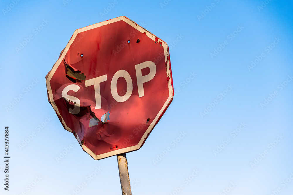 Damaged road sign stop covered with scratches and rusty. Rumpled road sign on a blue sky background. Stop sign with partly bent surface. Urban Grunge Background - obrazy, fototapety, plakaty 