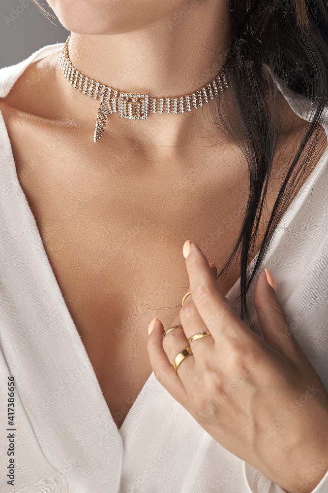 Cropped close-up shot of a lady holding her wrist near V-neckline of white blouse. The lady's neck is decorated with elegant bejeweled chocker with buckle. 