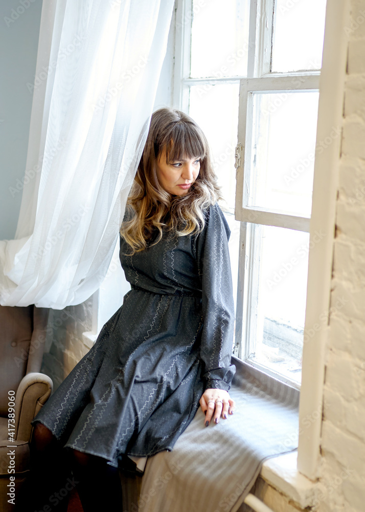 Young brunette woman in a dress sits dreamily by the window Young brunette woman in a dress sits dreamily by the window