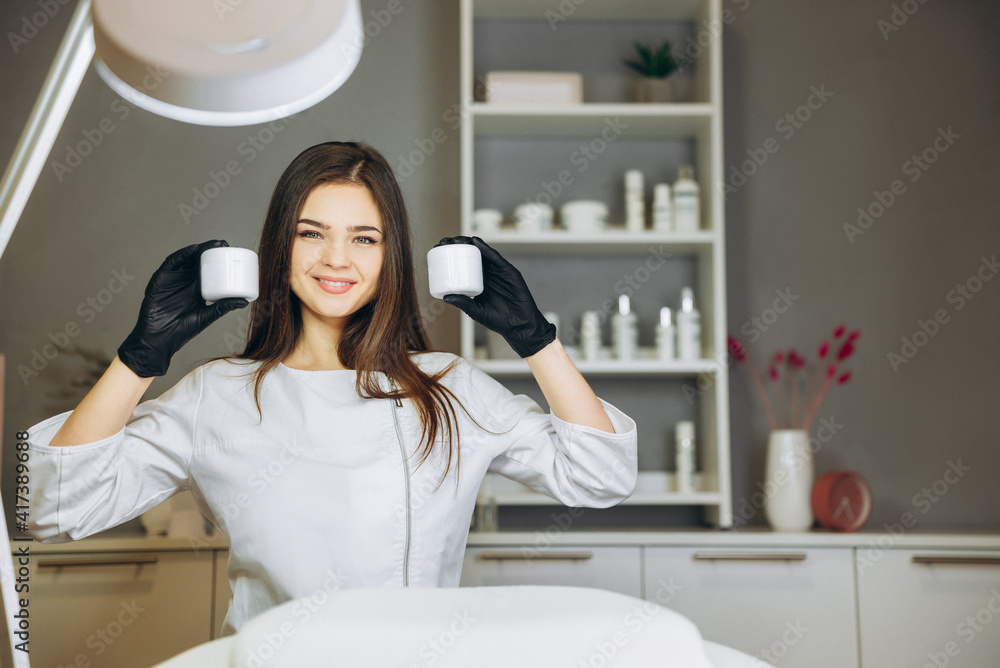 Consultation in cosmetology clinic. Female beauty doctor in white coat and black gloves show bottles of cream. Healthcare concept. Consultation in cosmetology clinic. Female beauty doctor in white coat and black gloves show bottles of cream. Healthcare concept.