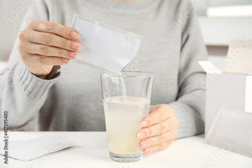 Woman pouring powder from medicine sachet into glass with water at table, closeup photo