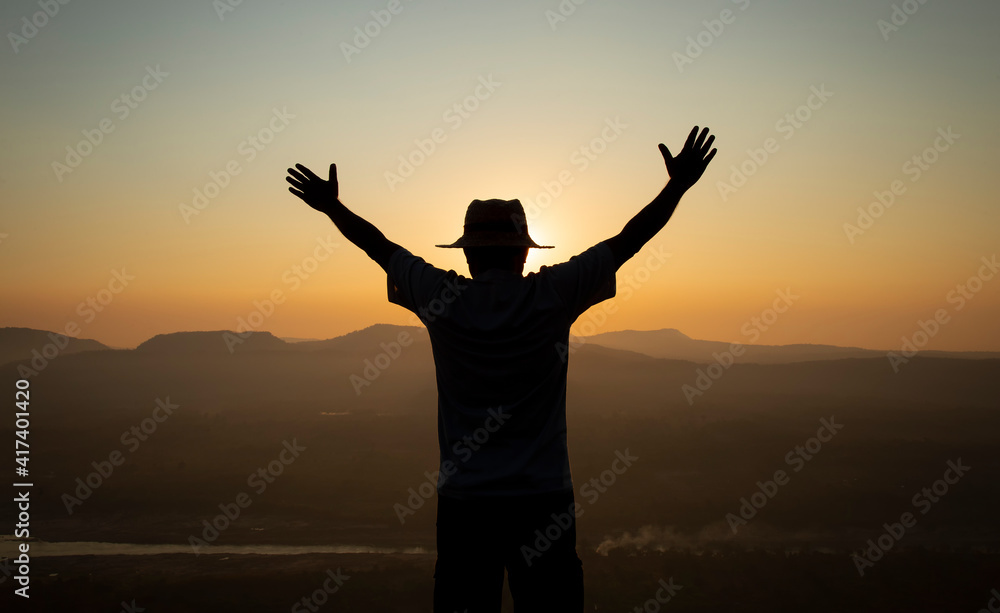 Successful silhouette hiker man standing raised hands on top of mountain, enjoying nature view during sunrise.