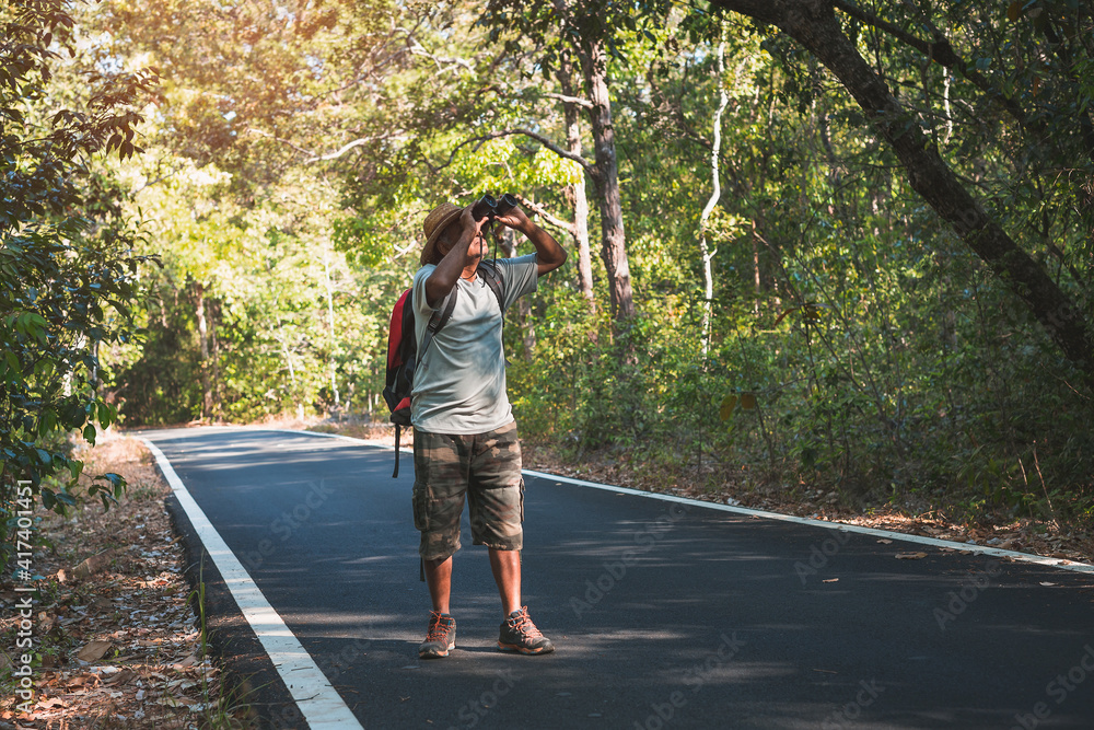 Fototapeta premium An elderly Asian man standing on asphalt road watching birds with binoculars on trees in the forest. Old man hiking in the vacation.