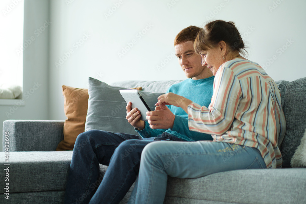 son teaching his mother to use tablet. older people using technology. Cheerful elderly woman sitting on the sofa next to his adult son son teaching his mother to use tablet. older people using technology. Cheerful elderly woman sitting on the sofa next to his adult son