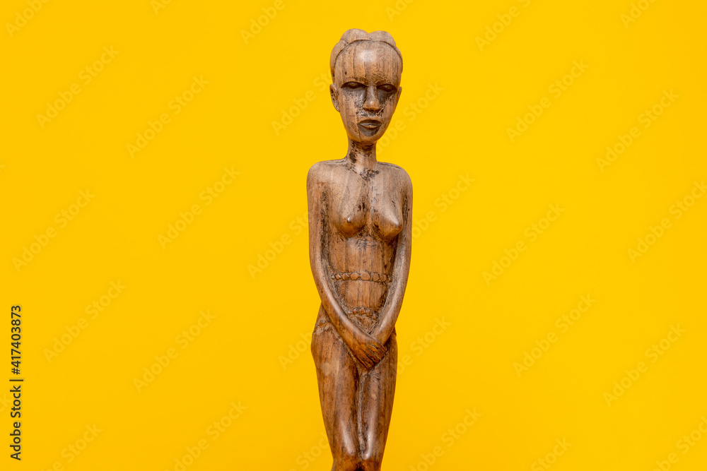 Upper part of a female figurine carved out of ebony wood without clothes on holding hands in front of her private area against a seamless yellow background. Upper part of a female figurine carved out of ebony wood without clothes on holding hands in front of her private area against a seamless yellow background.