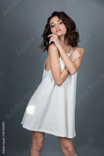studio portrait of a slender sexy long-legged brunette girl with a beautiful hairstyle in a white dress on a gray background in a photo studio © TetianaRUD