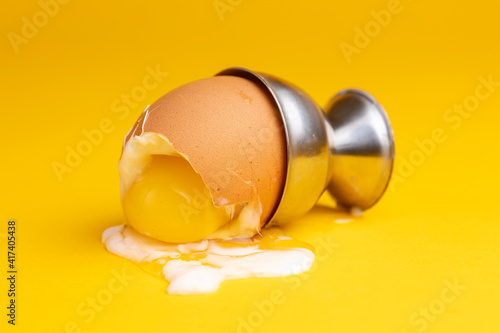 Photography Closeup of fallen over egg-cup with broken shell of soft cooked egg with the slimy white and yolk spilling over a seamless yellow background