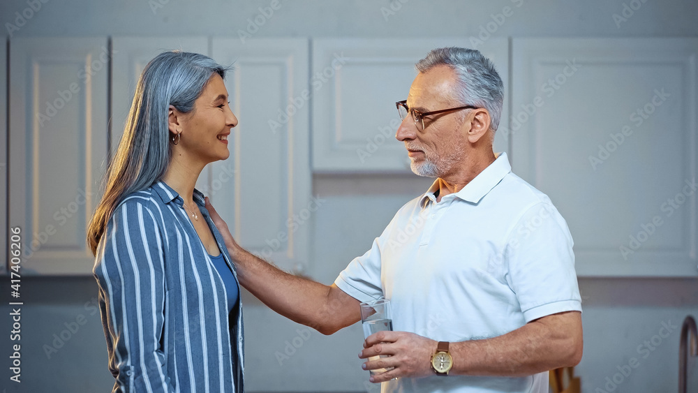 Fototapeta premium side view of smiling senior man holding glass of water while touching shoulder of happy asian wife