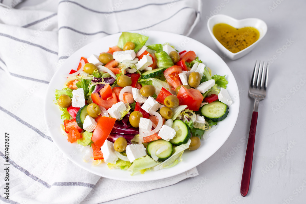 Delicious vegetable greek salad with feta, tomatoes, olives, greens on light background. Served with towel, fork and olive oil dressing. Healthy diet lunch idea. Quick, simple recipe.