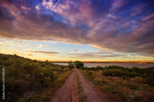 Rural road leading to the lake, river, at sunset, against the backdrop of beautiful clouds and the evening sky