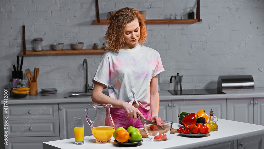 curly young woman cutting fresh tomato in kitchen curly young woman cutting fresh tomato in kitchen