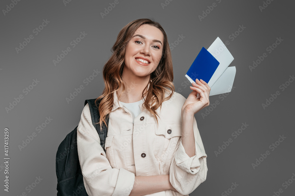 Student exchange concept. Happy Caucasian student girl holding foreign passport, tickets smiling isolated on gray background in studio Student exchange concept. Happy Caucasian student girl holding foreign passport, tickets smiling isolated on gray background in studio