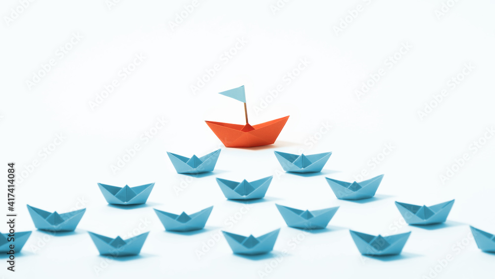 Orange paper boat ahead of a group of blue paper boats on white background. Concept of leadership, individuality, unique. Orange paper boat ahead of a group of blue paper boats on white background. Concept of leadership, individuality, unique.