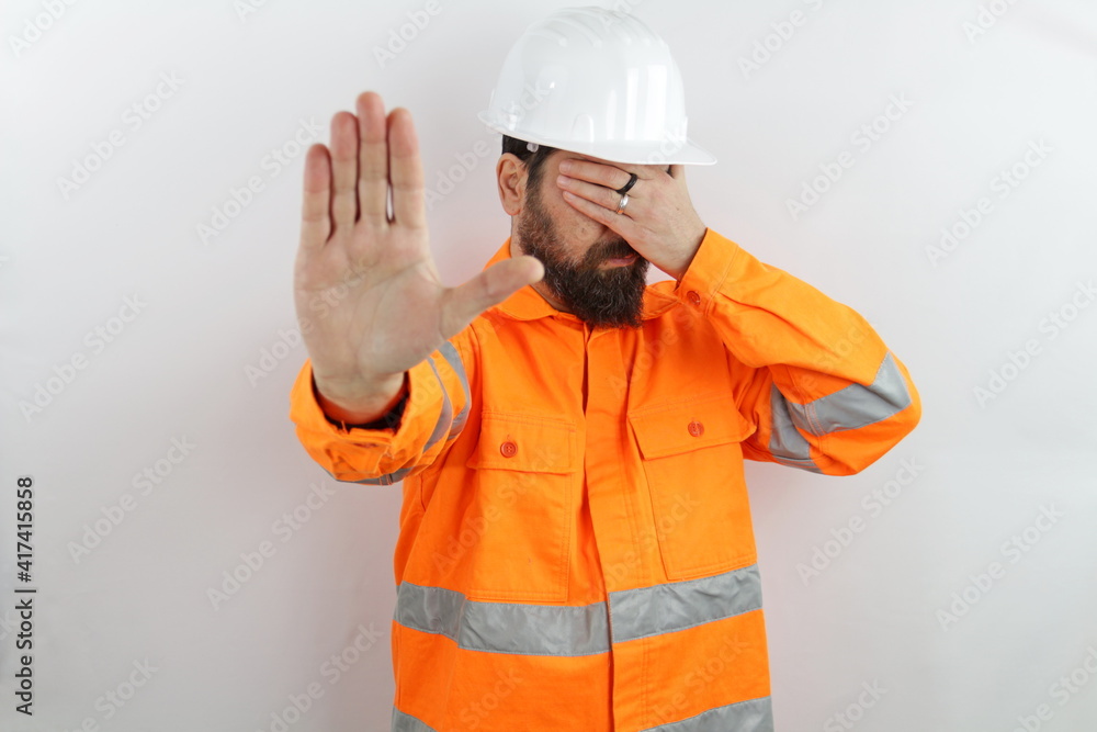 Bearded man wearing hardhat and reflecting jacket in shock covering face and eyes with hand with embarrassed expression and doing stop gesture with fear expression. Bearded man wearing hardhat and reflecting jacket in shock covering face and eyes with hand with embarrassed expression and doing stop gesture with fear expression.